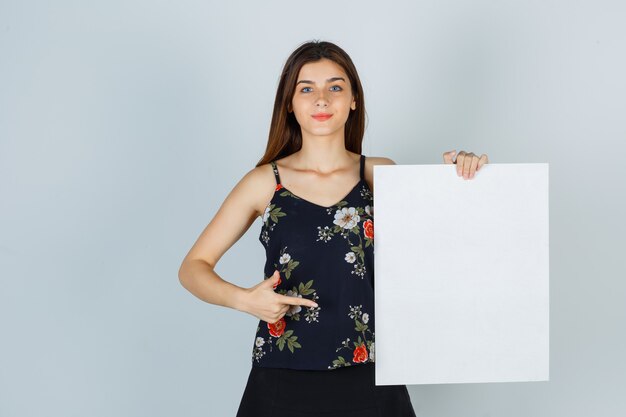 Jovem apontando para uma tela em branco na blusa, saia e olhando alegre. vista frontal.