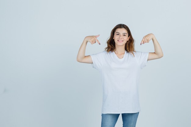 Jovem apontando-se em t-shirt, jeans e parecendo orgulhosa. vista frontal.