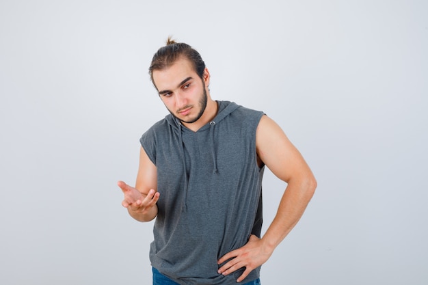 Jovem apto masculino com capuz sem mangas estendendo a mão em direção à câmera e olhando pensativo, vista frontal.