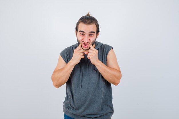 Jovem apto masculino com capuz sem mangas pressionando as bochechas com os dedos e parecendo feliz, vista frontal.