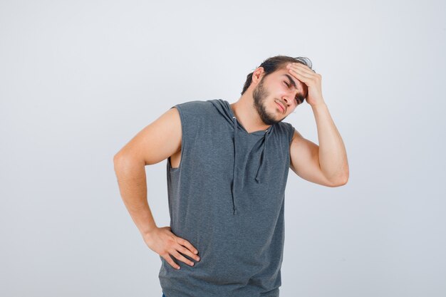 Jovem apto masculino, mantendo a mão na testa com um capuz sem mangas e parecendo dolorido, vista frontal.