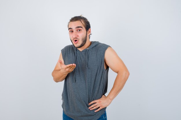 Jovem apto masculino posando com a mão na cintura enquanto espalha a palma da mão em um colete sem mangas e parecendo chocado. vista frontal.