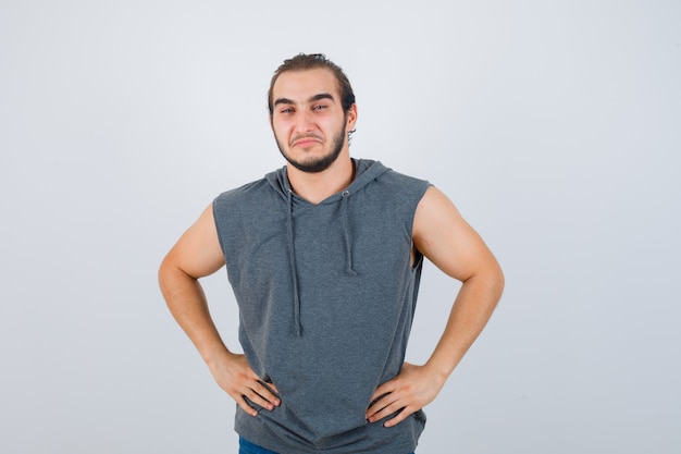 Foto grátis jovem apto masculino posando com as mãos na cintura em um capuz sem mangas e parecendo alegre. vista frontal.