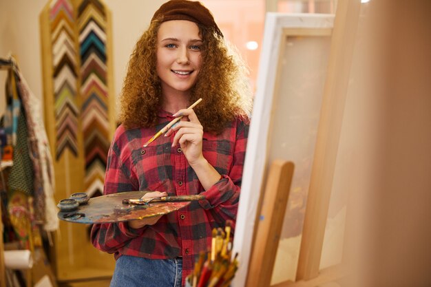 Jovem artista encaracolada parece feliz e segura uma paleta com tintas a óleo