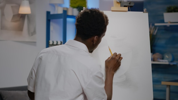 Foto grátis jovem artista negra retocando o projeto do vaso na tela e cavalete usando lápis e ferramentas de arte no espaço do estúdio de criatividade. afro-americano desenhando uma obra-prima profissional para hobby