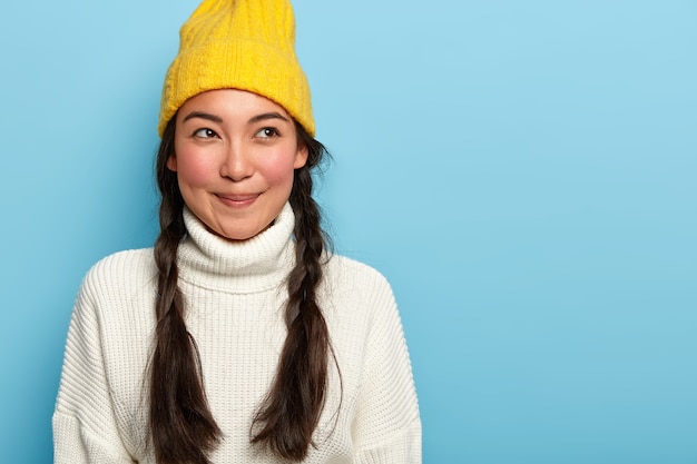 Jovem asiática atraente e pensativa com sorriso agradável, tem duas tranças escuras, vestida com roupa de inverno, isolada sobre fundo azul, expressa atitude encantadora