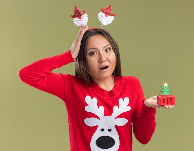 Jovem asiática com aro de cabelo de natal segurando um brinquedo de natal e colocando a mão atrás da cabeça isolada na parede verde oliva com medo