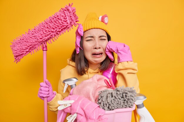 Jovem asiática descontente sente muito cansaço e chora porque não quer continuar limpando a casa segura o esfregão encostado na cesta com a roupa suja e os detergentes tem prazo para terminar de arrumar o quarto