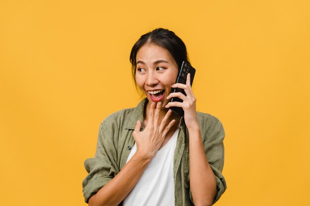 Jovem asiática fala por telefone com expressão positiva, sorri amplamente, vestida com roupas casuais, sentindo felicidade e fica isolado na parede amarela. Mulher feliz adorável feliz alegra sucesso.