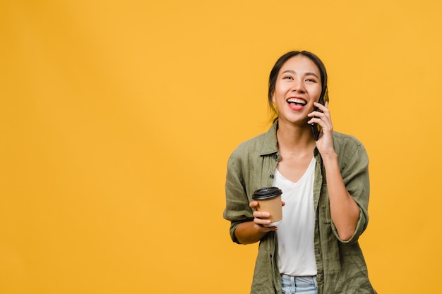 Jovem asiática falar por telefone e segurar a xícara de café com expressão positiva, sorrir amplamente, vestida com um pano casual, sentindo felicidade e ficar isolado na parede amarela. Conceito de expressão facial.