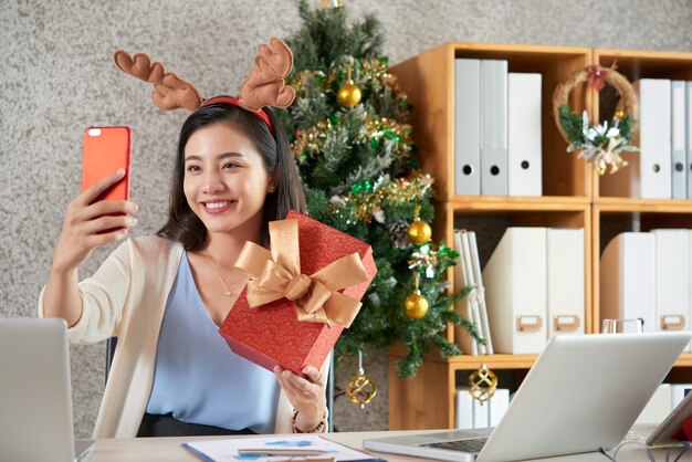 Jovem asiática feliz com uma fita de cabelo de rena tirando uma selfie com um presente de Natal