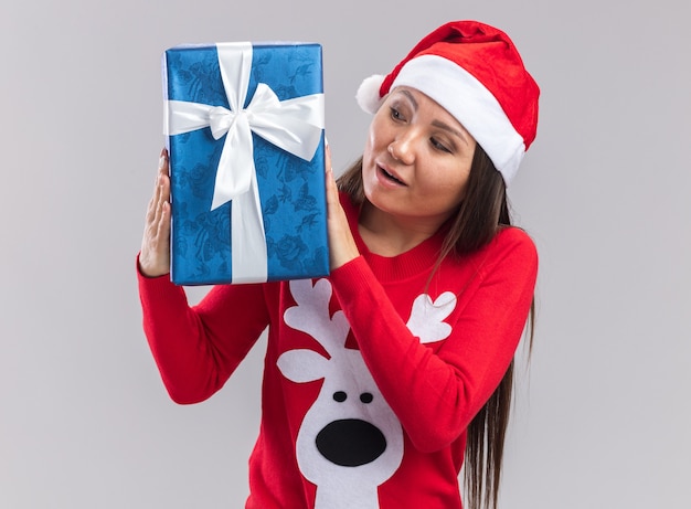Jovem asiática impressionada com chapéu de Natal e suéter segurando e olhando para uma caixa de presente isolada no fundo branco