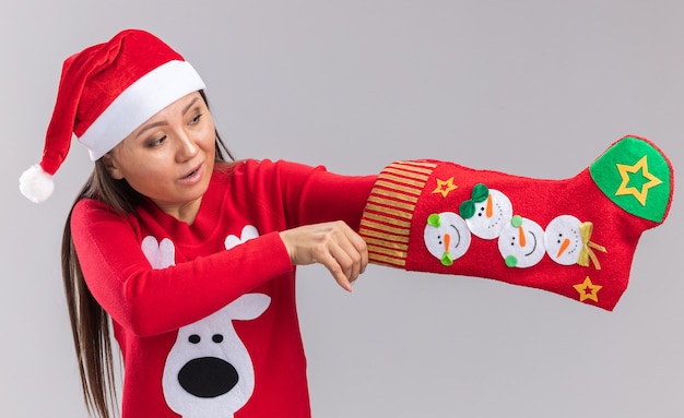 Jovem asiática impressionada usando chapéu de natal e suéter colocando a mão na meia de natal isolada na parede branca