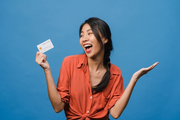 Jovem asiática mostra cartão de crédito com expressão positiva, sorri amplamente, vestida com roupas casuais, sentindo felicidade e estande isolado na parede azul. conceito de expressão facial.