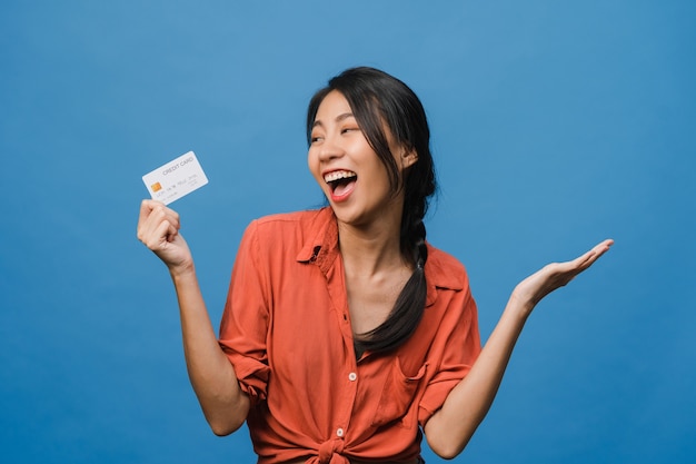 Jovem asiática mostra cartão de crédito com expressão positiva, sorri amplamente, vestida com roupas casuais, sentindo felicidade e estande isolado na parede azul. Conceito de expressão facial.