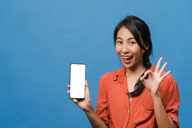 Jovem asiática mostra tela vazia do smartphone com expressão positiva, sorri amplamente, vestida com roupas casuais, sentindo felicidade na parede azul. Celular com tela branca na mão feminina.