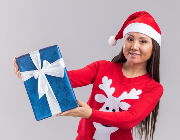 Jovem asiática sorridente usando chapéu de Natal e suéter segurando uma caixa de presente para a câmera