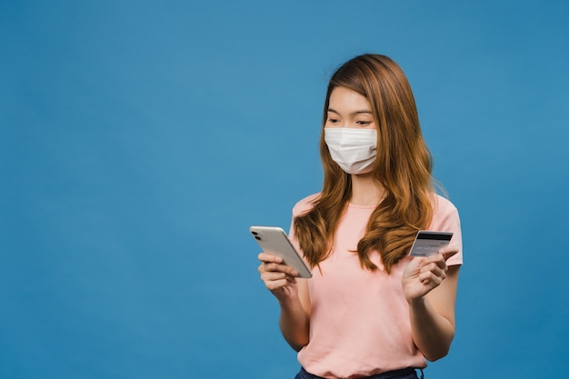 Foto grátis jovem asiática usando máscara médica usando telefone e cartão do banco de crédito com expressão positiva, sorri amplamente, vestida com roupas casuais e fica isolada na parede azul