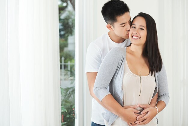 Jovem asiático em pé atrás de namorada, abraçando e beijando-a na bochecha