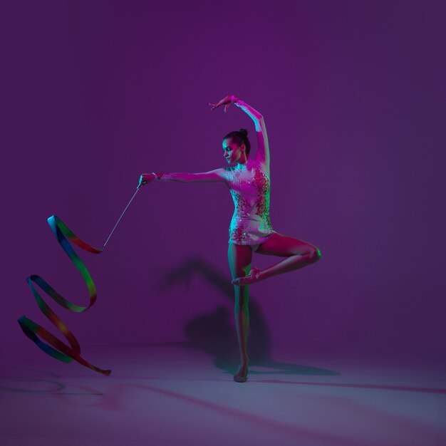 Jovem atleta, artista de ginástica rítmica dançando, treinando isolado no fundo roxo do estúdio com luz de néon. Linda garota praticando com o equipamento. Graça no desempenho.