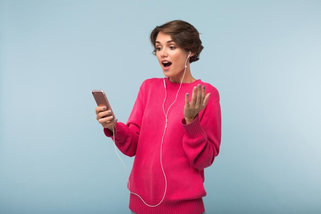 Jovem atônita de suéter rosa e fones de ouvido olhando espantada no celular sobre fundo azul isolado