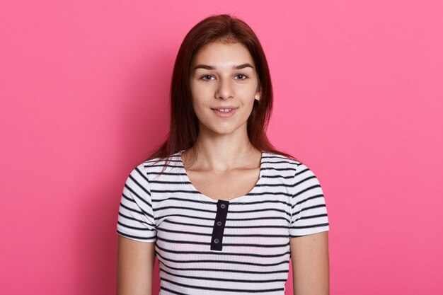 Jovem atraente adolescente vestindo camiseta listrada casual, posando contra uma parede rosa, com um sorriso encantador, parece feliz.