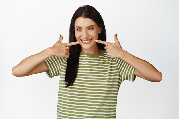 Jovem atraente apontando dedos para seu sorriso mostrando dentes brancos e saudáveis em pé sobre fundo branco em camiseta