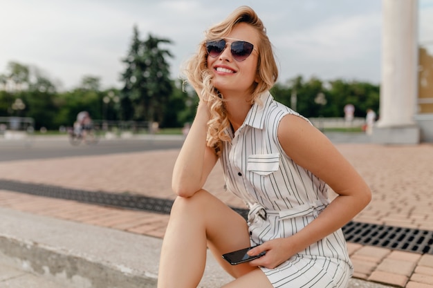 Jovem atraente bonita loira elegante sentada na rua da cidade com vestido de algodão branco estilo moda de verão usando óculos escuros