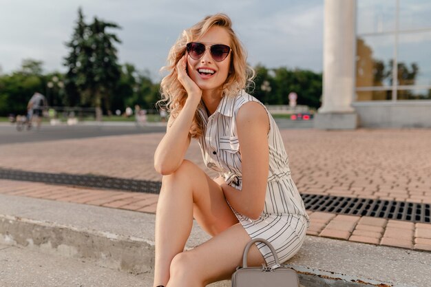 Jovem atraente bonita loira elegante sentada na rua da cidade com vestido de algodão branco estilo moda de verão usando óculos escuros