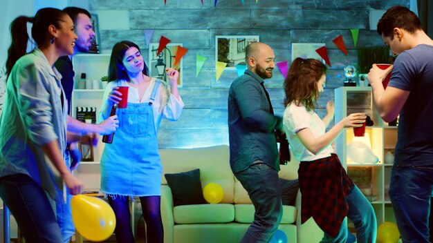 Jovem atraente careca sorrindo enquanto dançava na festa com seus amigos. Festa universitária selvagem com luzes de néon e bola de discoteca