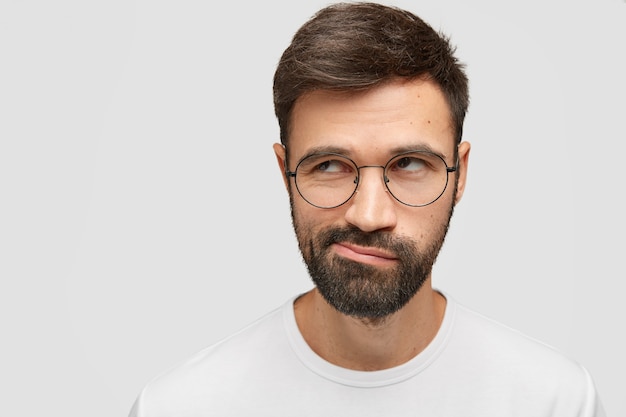 jovem atraente com a barba por fazer parece duvidoso, pensativamente de lado, franze os lábios, tem uma barba espessa e escura