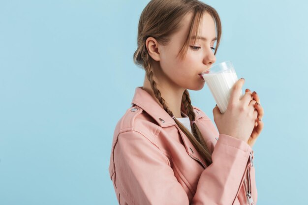 Jovem atraente com duas tranças na jaqueta de couro rosa sonhadoramente bebendo leite enquanto passa o tempo sobre fundo azul