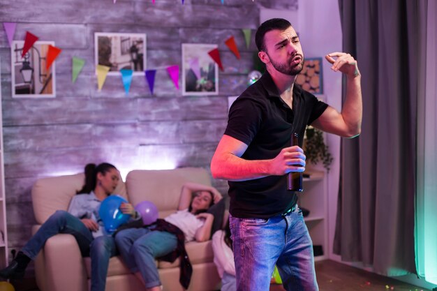 Jovem atraente dançando uma garrafa de cerveja na mão em uma festa enquanto seus amigos estão dormindo.