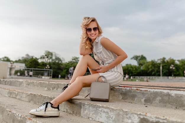 Jovem atraente e elegante mulher loira sentada na rua da cidade, com vestido de estilo de moda de verão, usando óculos escuros, bolsa e tênis prateados