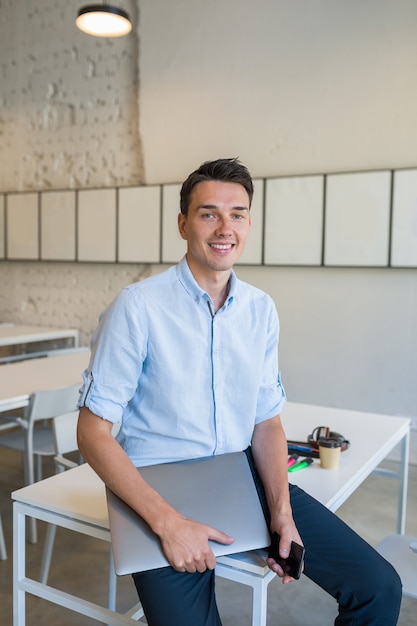 Jovem atraente e sorridente, sentado em um escritório aberto em colaboração, segurando um laptop