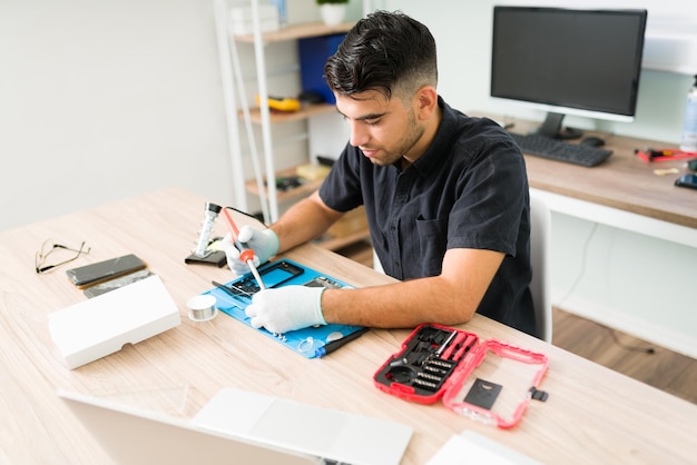 Jovem atraente e técnico usando um ferro de solda enquanto tenta consertar o hardware de um smartphone danificado