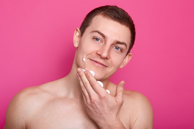 Jovem atraente feliz limpa gel de barbear do rosto, parece satisfeito. O modelo de olhos azuis atlético nu posa tocando o rosto com uma mão, olhando para o outro lado. Copie o espaço para propaganda.