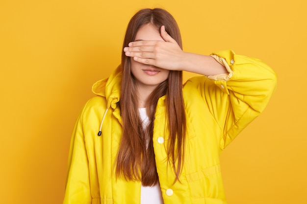 Jovem atraente feminino vestindo jaqueta cobrindo os olhos com as mãos, mulher com cabelos longos, de pé contra a parede amarela, senhora se esconde de sua amiga.
