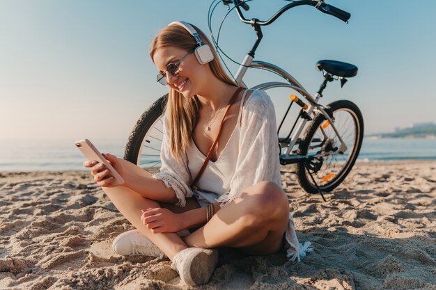 Jovem atraente loira sorridente, elegante, sentada na praia com uma bicicleta em fones de ouvido, ouvindo música