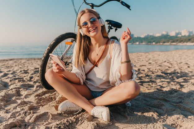 Jovem atraente loira sorridente, elegante, sentada na praia com uma bicicleta em fones de ouvido, ouvindo música