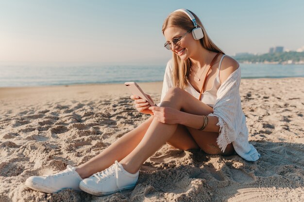 Jovem atraente loira sorridente, elegante, sentada na praia com uma bicicleta em fones de ouvido, ouvindo música