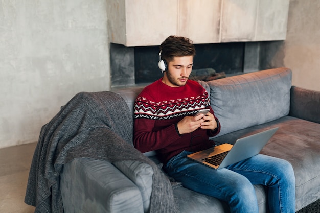 Jovem atraente no sofá em casa no inverno com smartphone em fones de ouvido, ouvindo música, vestindo uma blusa de malha vermelha, trabalhando no laptop, freelancer