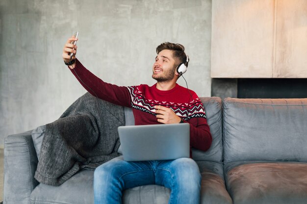 Jovem atraente no sofá em casa no inverno com smartphone em fones de ouvido, ouvindo música, vestindo uma blusa de malha vermelha, trabalhando no laptop, freelancer