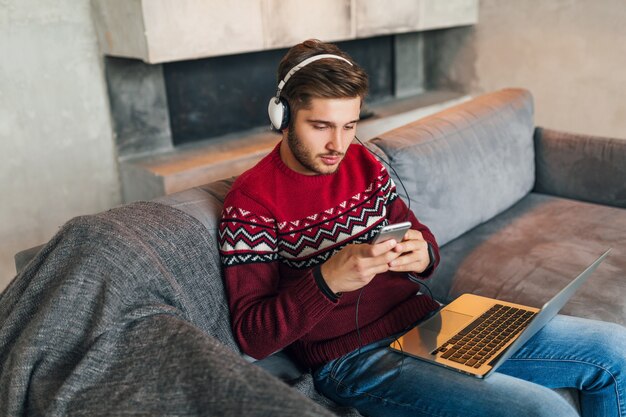 Jovem atraente no sofá em casa no inverno com smartphone em fones de ouvido, ouvindo música, vestindo uma camisola de malha vermelha, trabalhando no laptop, freelancer