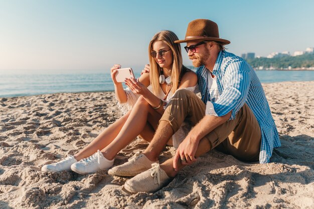 Jovem atraente sorridente, feliz, homem e mulher com óculos de sol, sentado na praia de areia tirando foto de selfie