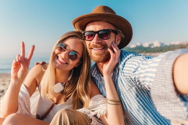 Jovem atraente sorridente, feliz, homem e mulher com óculos de sol, sentado na praia de areia tirando foto de selfie