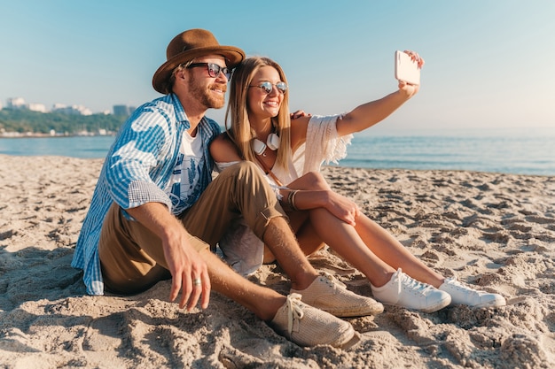 Jovem atraente sorridente, feliz, homem e mulher em óculos de sol, sentado na praia de areia, tirando foto de selfie