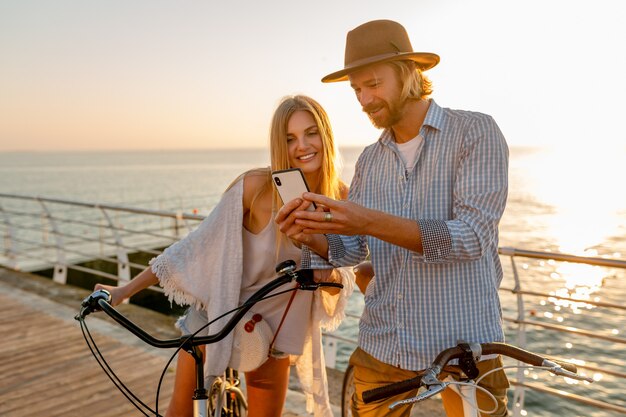 Jovem atraente sorridente feliz homem e mulher viajando de bicicleta usando smartphone, casal romântico à beira-mar no pôr do sol, roupa de estilo boho hipster, amigos se divertindo juntos