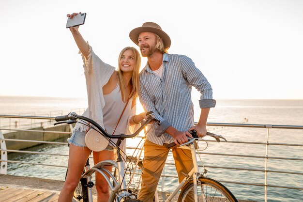 Jovem atraente sorridente homem feliz e mulher viajando de bicicleta tirando foto de selfie na câmera do telefone, casal romântico à beira-mar no pôr do sol, roupa estilo boho hipster, amigos se divertindo juntos