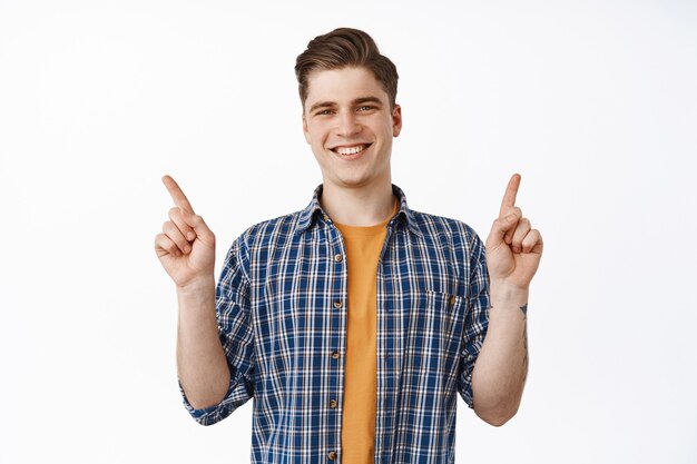 Jovem atraente sorridente, universitário apontando os dedos de lado a variedade de descontos e parece feliz, em pé sobre o branco.